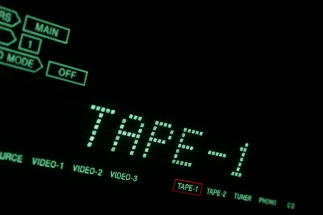 Green Hi-Fi LCD display in close-up TAPE, A hi-fi receiver's green dot-matrix LCD display in extreme close-up showing TAPE-1 (cassette). Tilted view with green font on black background.