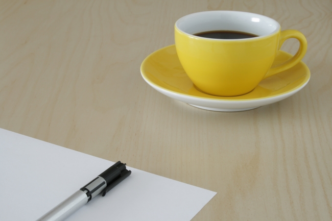 Yellow cup of coffee on wooden table, sheet of paper and pen, Yellow cup of coffe on a wooden modern office table. Cup in upper right corner, in the foreground, the lower left, a piece of white paper and a silver and bl...