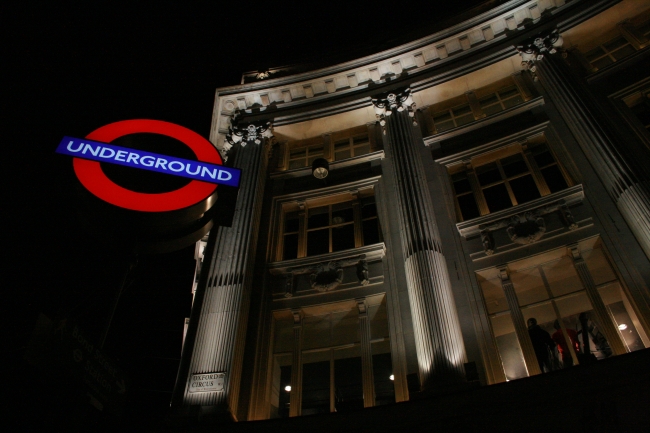Oxford Circus Underground sign at night, with an illuminated fascade behind it