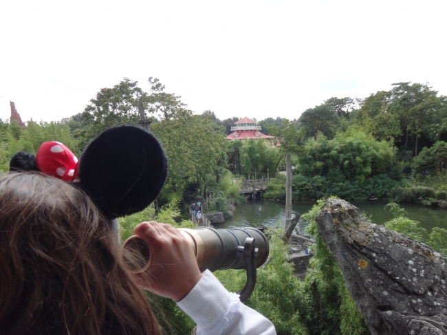 A little girl with Minnie Mouse ears, looking through the spyglass from the lookout over Adventure Isle lake and Col. Hathi's in the background
